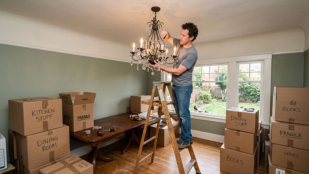 Removing Fixtures Before Selling: What Stays and What Goes? A homeowner standing on a stepladder carefully unscrewing an ornate dining room chandelier, surrounded by packed cardboard moving boxes.